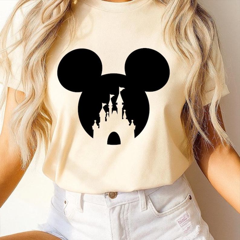 Puede incluir: Camiseta color crema con una silueta negra de la cabeza de Mickey Mouse y un dise&ntilde;o de castillo en su interior. La camiseta tiene mangas cortas y cuello redondo. El dise&ntilde;o est&aacute; centrado en la parte delantera.