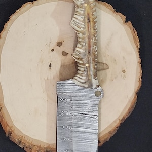 May include: A handmade knife with a patterned steel blade and a handle made of bone-like material. The knife has a ring at the top and is displayed on a wooden slice.