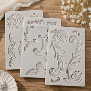 May include: Three rectangular white silicone molds featuring intricate embossed patterns. The designs include floral motifs, scrolling acanthus leaf flourishes, and a small winged cherub figure, all recessed into the flexible material surface.