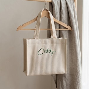 Peut inclure: Un sac fourre-tout en toile beige à rayures verticales et à courtes poignées, suspendu à un cintre en bois. Le sac porte le mot "Cataleya" en écriture verte. Un tissu gris est suspendu derrière le sac.
