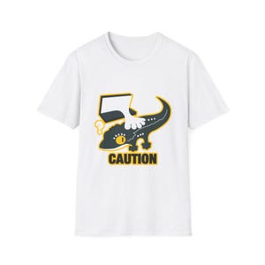 Könnte beinhalten: Weißes T-Shirt mit einer Cartoon-Illustration einer dunkelgrünen Eidechse, auf die ein weißer Fuß tritt. Das Wort "CAUTION" steht in gelben Blockbuchstaben unter der Eidechse.