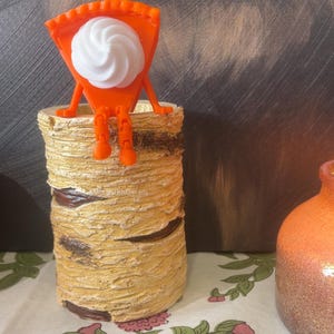 May include: A bright orange pie slice figurine with a white whipped cream dollop and articulated orange limbs, perched on the rim of a cylindrical container textured to resemble a piece of birch tree bark with horizontal brown markings.
