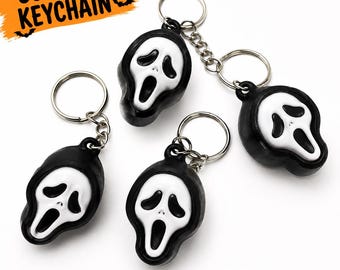 Puede incluir: Tres llaveros con una máscara blanca de estilo fantasma con aberturas negras para los ojos y la boca, unidos a una anilla de metal plateado. La máscara tiene un borde negro. El texto dice SCREAM KEYCHAIN y CUTE SPOOKY COLLECTIBLE.
