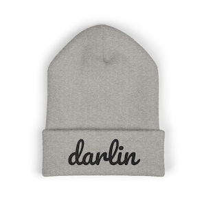 May include: A light gray beanie hat with the word "darlin" in black script across the folded brim. The hat is made of a soft, knitted material.
