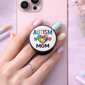 May include: A round phone grip with the words "AUTISM MOM" and a colorful puzzle heart design. The grip is black and white, attached to a pink phone. The phone has a silver ring and is held by a person with painted nails.