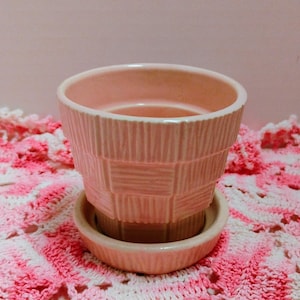 Könnte beinhalten: Hellrosa Keramik-Blumentopf mit passender Untertasse. Der Topf hat ein strukturiertes Korbgeflecht-Design. Der Topf steht auf einer gehäkelten Oberfläche in Rosa und Weiß.