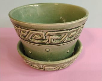 May include: A green ceramic planter with a matching saucer. The planter has a decorative band of repeating geometric patterns around the top and the saucer. The planter is approximately 15 cm in diameter.