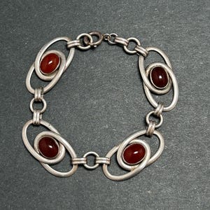 May include: A silver-toned metal bracelet featuring four oval links, each containing a smooth, dark red, oval-shaped stone. The links are connected by small circular rings, and the bracelet includes a circular spring-ring clasp for closure.