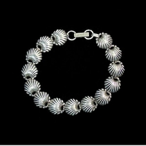 May include: Silver bracelet featuring a series of seashell-shaped links. The bracelet has a secure clasp and is displayed against a black background. The seashell links are detailed with textured ridges, creating a nautical theme.