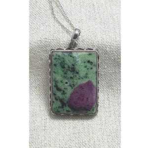 May include: A rectangular pendant necklace featuring a green and black stone with a large, deep red section. The stone is set in a silver-colored metal frame and hangs from a delicate silver chain. The pendant is a unique piece of jewelry.