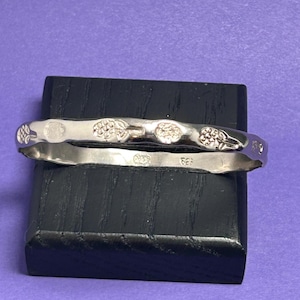 May include: A silver-colored metal bangle bracelet featuring a repeating embossed pattern of small floral or fruit-like motifs along the outer surface. The interior of the band is stamped with the text 925, indicating sterling silver composition.