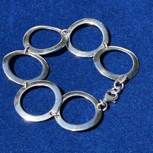 May include: A silver bracelet featuring six interconnected oval links. The bracelet is made of polished metal and has a secure clasp. The jewelry is displayed on a blue surface.