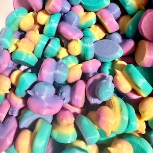May include: A close-up of a pile of colorful, 3D-printed objects in various shapes, including ducks, hearts, and bowties. The objects are in pastel colors, including yellow, teal, purple, and pink. The objects are likely toys or decorative items.