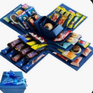 Op de afbeelding: Een blauwe explosiedoos gevuld met diverse verpakte snoepjes en lekkernijen. De doos is open en onthult meerdere lagen lekkernijen, met een kleinere doos en een blauwe strik aan de onderkant. De lekkernijen zitten in kleurrijke verpakkingen.