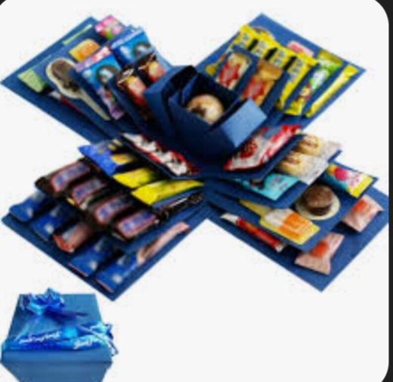 Op de afbeelding: Een blauwe explosiedoos gevuld met diverse verpakte snoepjes en lekkernijen. De doos is open en onthult meerdere lagen lekkernijen, met een kleinere doos en een blauwe strik aan de onderkant. De lekkernijen zitten in kleurrijke verpakkingen.