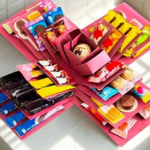 Op de afbeelding: Een roze explosiedoos, opengeklapt om lagen verpakte snoepjes, chocolaatjes en kleine traktaties te onthullen. De doos is in een stervormig patroon gerangschikt, met de inhoud zichtbaar. De traktaties zitten in kleurrijke verpakkingen.