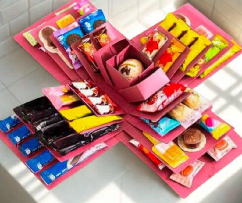 Op de afbeelding: Een roze explosiedoos, opengeklapt om lagen verpakte snoepjes, chocolaatjes en kleine traktaties te onthullen. De doos is in een stervormig patroon gerangschikt, met de inhoud zichtbaar. De traktaties zitten in kleurrijke verpakkingen.