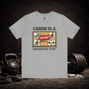 Puede incluir: Camiseta gris con la frase "Cardio Is A Government Plot". La camiseta presenta un gráfico humorístico de equipos de ejercicio y un hombre de palitos, con una etiqueta "Top Secret". Una pesa rusa y pesas están en el fondo.