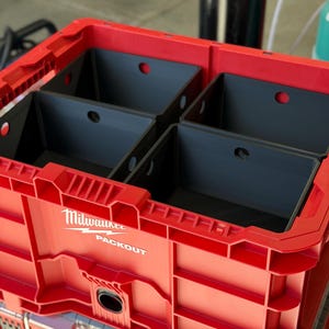 May include: A red Milwaukee Packout storage crate featuring four black rectangular plastic bins inserted inside. The crate has a rigid, modular design with visible branding text that reads Milwaukee Packout on the front exterior panel.