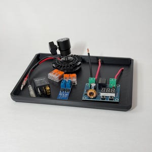 May include: A black rectangular tray containing electronic components including a Bosch relay, orange wire connectors, a small blue circuit board, and a larger blue circuit board with a digital display and a copper coil, plus a black camera mount.