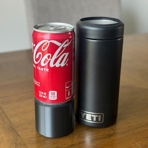 May include: A red 7.5 fluid ounce Coca-Cola can sitting inside a black cylindrical plastic base, positioned next to a black YETI brand insulated can cooler. The can features white text and branding, while the cooler has a silver YETI logo.
