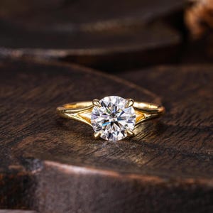 May include: A gold ring featuring a single round brilliant-cut clear gemstone held in a four-prong setting. The band has a smooth, polished finish and splits into a V-shape where it meets the central stone, creating a clean and structured look.