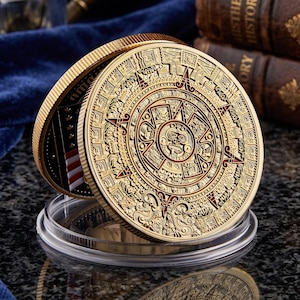 May include: A gold-coloured circular coin featuring an intricate Aztec calendar design with red enamel accents. The coin is displayed upright in a clear plastic protective case, revealing a partial view of an American flag on its reverse side.