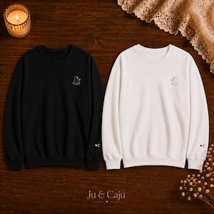 Könnte beinhalten: Ein schwarzes Sweatshirt und ein weißes Sweatshirt, jeweils mit einer kleinen gestickten Geistergrafik auf der linken Brust und einem kleinen Herz mit dem Buchstaben C am linken Ärmelbündchen. Beide Kleidungsstücke haben einen Rundhalsausschnitt.