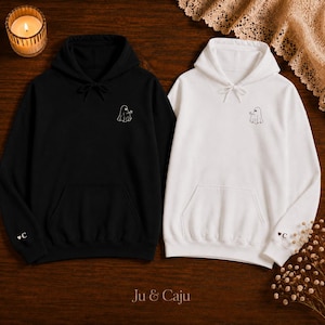 Könnte beinhalten: Ein schwarzer Kapuzenpullover und ein weißer Kapuzenpullover, jeweils mit einem kleinen gestickten Geist auf der Brust und einem Herz mit dem Buchstaben C am Ärmelbündchen. Beide Pullover haben eine Kapuze mit Kordelzug und eine Kängurutasche.