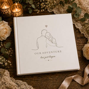 May include: A square white photo album with a minimalist line art illustration of a couple embracing. Above the figures is a small heart. Below the illustration, the text reads "OUR ADVENTURE has just begun" with a small heart and line detail.