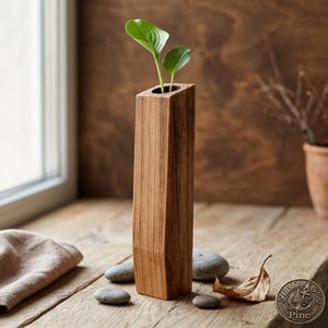 Könnte beinhalten: Eine hohe, geometrische Holzvase mit quadratischer Öffnung, die zwei grünblättrige Pflanzen enthält. Die Vase besteht aus hellbraunem Holz und steht auf einer Holzoberfläche mit kleinen Steinen und einem getrockneten Blatt. Die Vase ist etwa 30 cm hoch.