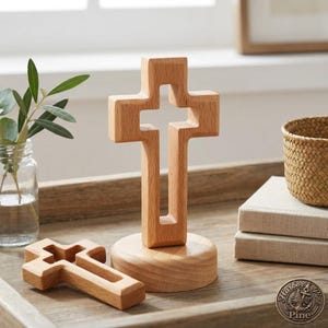 Puede incluir: Dos cruces de madera clara, una de pie sobre una base redonda y otra tumbada. Ambas presentan un diseño calado. También se ve un pequeño tarro de cristal con hojas verdes.