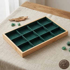 Könnte beinhalten: Ein rechteckiges Schmucktablett aus Holz mit einem grünen Filzinnenraum, der in zwölf Fächer unterteilt ist. Das Tablett steht auf einer neutralfarbenen Leinenoberfläche, mit kleinen grünen Edelsteinen und einem Stück Treibholz.