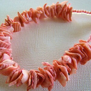May include: A pink shell necklace made of small, flat, and irregularly shaped shells. The necklace is strung on a thin, white cord.