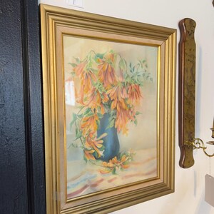 Puede incluir: Pintura enmarcada de un bodegón floral. La obra presenta flores naranjas en un jarrón azul, sobre un fondo suave y neutro. El marco es dorado, lo que añade un toque clásico a la pieza. La pintura se exhibe en una pared.