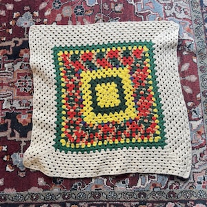 May include: A square, handmade crochet blanket with a vibrant, colorful design. The central square features a pattern of yellow, red, and green, framed by a green border and a cream-colored outer edge. The blanket is made with a granny square pattern.