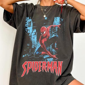May include: Dark gray t-shirt featuring a red and blue Spider-Man graphic. The design includes the word "SPIDERMAN" in a bold, red font. The shirt has a vintage, worn look.