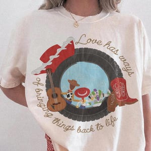 K&ouml;nnte beinhalten: Ein elfenbeinfarbenes Kurzarm-T-Shirt mit einer Grafik, die einen Reifen zeigt, der Zeichentrickfiguren, eine Gitarre, einen roten Hut und einen roten Cowboystiefel umrahmt. Der Text lautet, Love has ways of bringing things back to life.