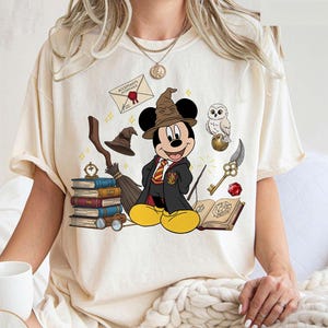 May include: Ivory-colored t-shirt featuring a cartoon Mickey Mouse dressed as a wizard, with a brown hat and black robe. The design includes books, a broom, an owl, and a letter. The word "IVORY" is printed at the bottom.