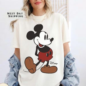 May include: Ivory-colored t-shirt featuring a large graphic of Mickey Mouse in his classic pose. Mickey is depicted in black, red, and brown. The word "Disney" is printed below the character. The shirt also has the text "NEXT DAY SHIPPING".