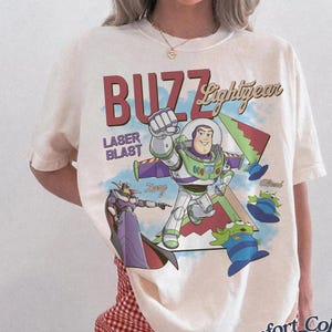 May include: Ivory t-shirt with a graphic of Buzz Lightyear from Toy Story. The design includes the words "Buzz Lightyear" and "Laser Blast," along with images of Zurg and the aliens. The shirt is a Comfort Colors brand.