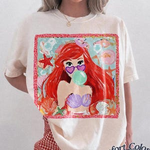 May include: Ivory-colored t-shirt featuring a colorful graphic of a mermaid with red hair, heart-shaped sunglasses, and a bubble. The graphic is surrounded by seashells and starfish. The shirt is labeled "Comfort Colors Ivory".