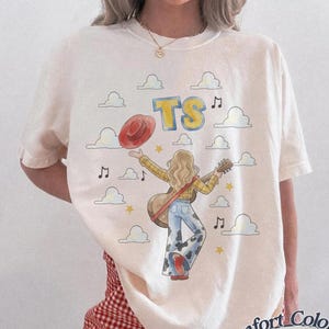 May include: An ivory short-sleeve t-shirt featuring a graphic illustration of a person with long blonde hair playing an acoustic guitar, wearing a yellow plaid shirt and cow-print pants, with the text TS, musical notes, and clouds.