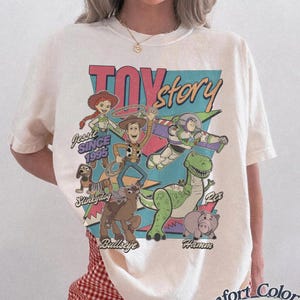 May include: Ivory-coloured t-shirt with a vibrant graphic of Toy Story characters, such as Woody and Buzz Lightyear. The design includes the text "Toy Story" and "Since 1995." The shirt is from Comfort Colors.