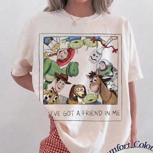 May include: Ivory-colored t-shirt featuring a rectangular graphic of Toy Story characters, including Woody, Buzz Lightyear, and Rex. The text "I'VE GOT A FRIEND IN ME" is below the graphic. The shirt is a Comfort Colors brand.