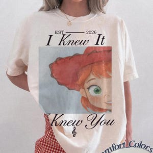 May include: An ivory short-sleeve t-shirt featuring a graphic of a cartoon character with red hair and a red hat. The text reads EST 2026 I Knew It Knew You with a treble clef symbol. The shirt is paired with red and white checkered shorts.