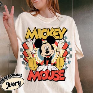 May include: Ivory-colored t-shirt featuring a graphic of Mickey Mouse. The design includes the words "Mickey Mouse" in yellow and red, with Mickey in the center. The shirt has a vintage, distressed look, with a "Comfort Colors" label.