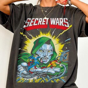 Puede incluir: Camiseta gris oscuro con una ilustraci&oacute;n de c&oacute;mic y el texto "SECRET WARS". El gr&aacute;fico representa una figura con capucha verde con una cara met&aacute;lica, rodeada de personajes de c&oacute;mic. La camiseta tiene un aspecto vintage.