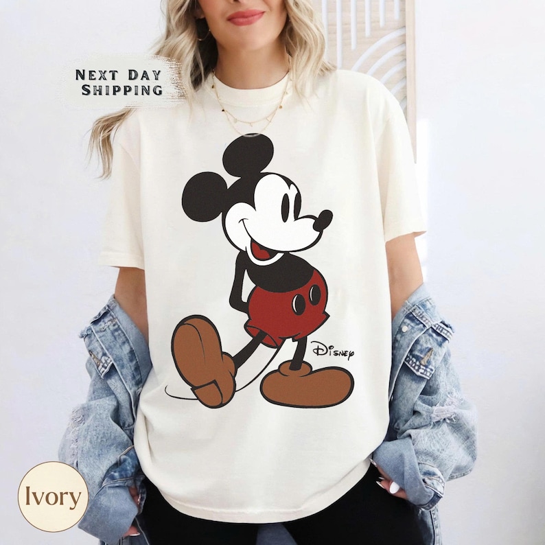May include: Ivory-colored t-shirt featuring a large graphic of Mickey Mouse in his classic pose. Mickey is depicted in black, red, and brown. The word "Disney" is printed below the character. The shirt also has the text "NEXT DAY SHIPPING".