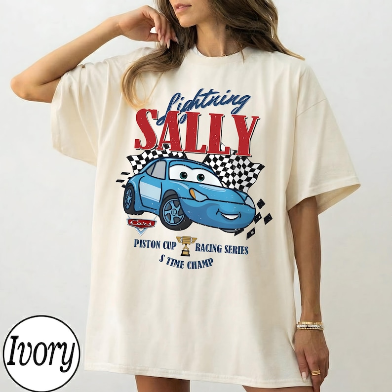 May include: Ivory-coloured t-shirt featuring a blue cartoon car with the text "Lightning Sally" in red and blue. The design includes a checkered flag and the words "Piston Cup Racing Series 5 Time Champ."